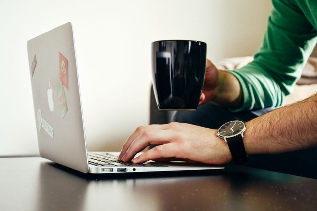 Individual programming on laptop while holding coffee mug with stickers, wearing wristwatch, casual green sweater. Suitable for depicting freelance work, remote working environments, home office setups, or lifestyle blogging. Use in articles about productivity, casual work environments, or tech-driven industries.