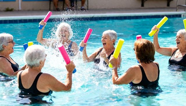 Senior women enjoying water aerobics class, splashing with colorful foam noodles