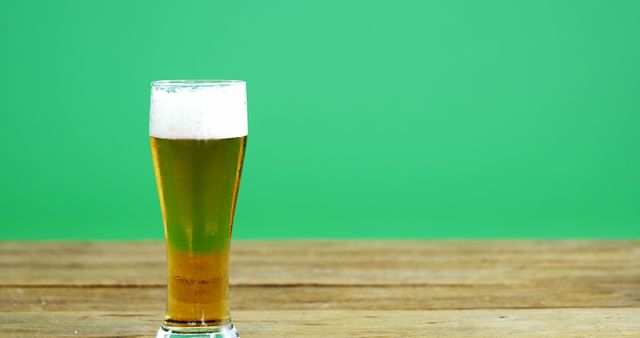 Tall glass of golden beer with thick frothy head placed on wooden table with vibrant green background. Perfect for advertising bars, pubs, or breweries, and promotional materials for drink-related events.