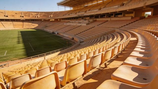Sunlit curved stadium seats forming rhythmic shadows over green pitch and empty tiers