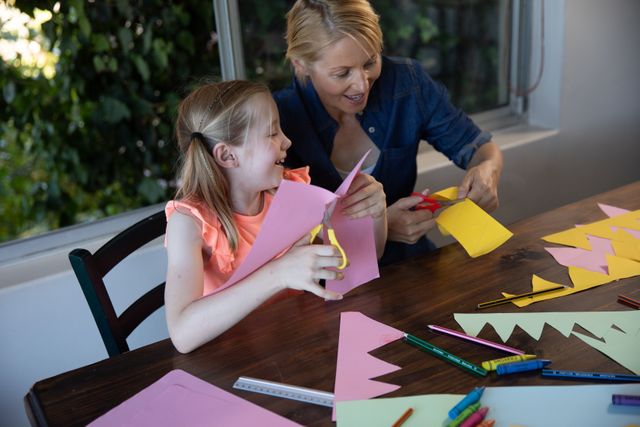 Caucasian woman spending family time together with her daughter at home, sitting at table in dining room cutting with scissors creative art. Family leisure time at home.