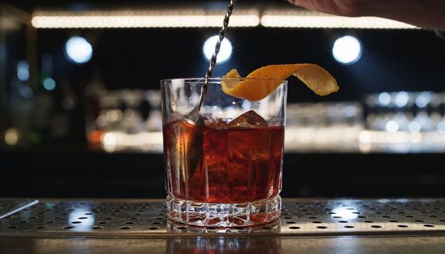 Stirring negroni cocktail in crystal rocks glass with orange twist, moody bar bokeh