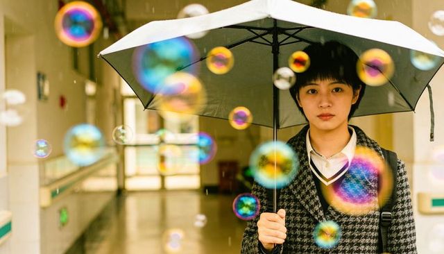 Asian teen holding umbrella amid colorful floating bubbles in campus hall dreamy portrait