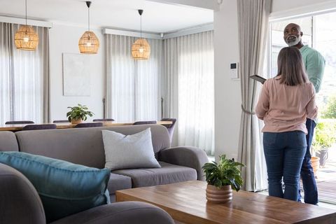 Senior couple standing by sliding glass door holding tablet in bright modern living room