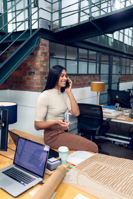 Smiling biracial female entrepreneur talking on mobile phone while sitting on desk in modern office. Ideal for use in business, entrepreneurship, technology, and workplace productivity themes. Perfect for illustrating modern office environments, professional communication, and creative workspaces.