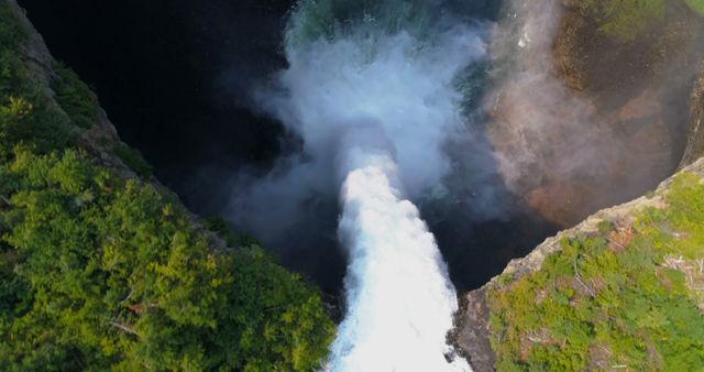 Aerial view captures the powerful force of a waterfall cascading into a natural pool surrounded by lush greenery. The mist from the waterfall creates a mystical atmosphere as it blends with the surrounding environment.