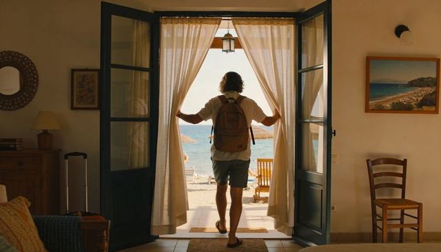 Traveler stepping through sunlit doorway opening onto mediterranean beach with backpack