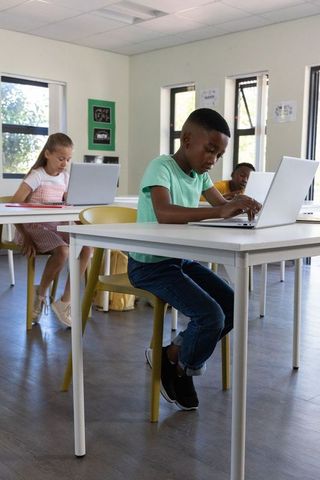 Young african american boy working on laptop in bright classroom with classmates