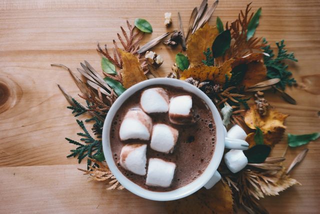 Hot cocoa topped with marshmallows on a wooden table, surrounded by autumn leaves and pinecones. Perfect for cozy autumn and winter themes, beverage advertisements, coffee shop decor, or seasonal greeting cards.