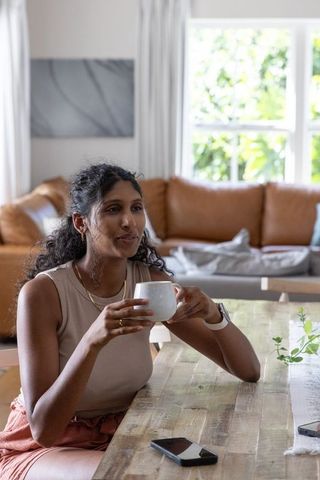 Sunlit african american woman relaxing at wood table holding coffee mug, casual home