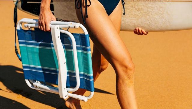 Woman carrying foldable beach chair and surfboard on sunlit sand walking to shore