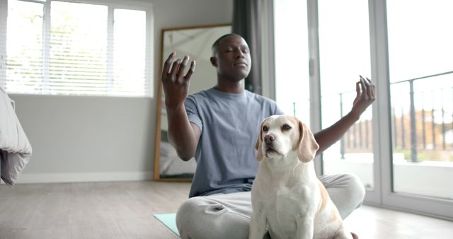 African american man practicing yoga meditation sitting on mat at home with pet dog. Healthy lifestyle, wellbeing, free time, pets and domestic life, unaltered.