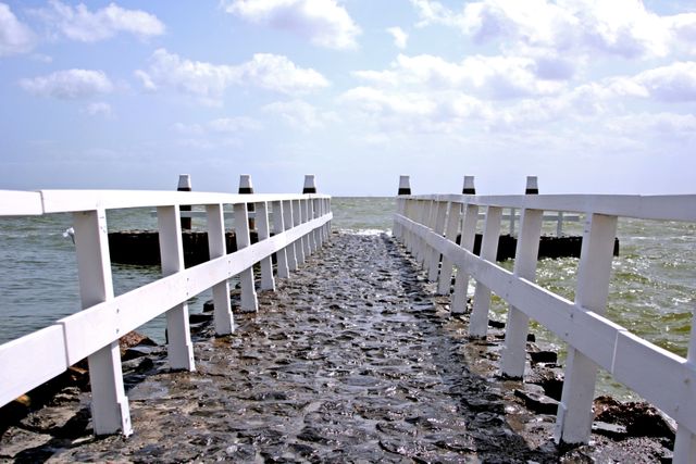 Pier extending into calm ocean with cobblestone pathway and white railings framed by a cloudy sky and a serene seascape. Ideal for travel brochures, inspirational quotes, calming backgrounds, and coastal vacation advertisements.