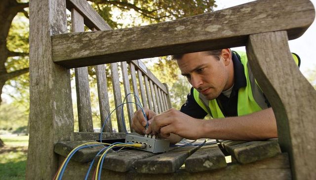 Technician repairing outdoor telecom cables on park bench wearing hi-vis vest