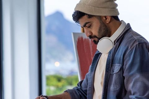 Contemporary bearded artist painting red canvas wearing beanie and headphones by large window