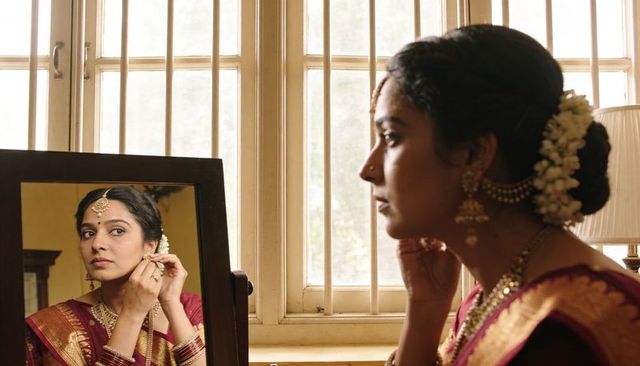 Indian bride adjusting earring in mirror wearing maroon silk sari and gold temple jewelry