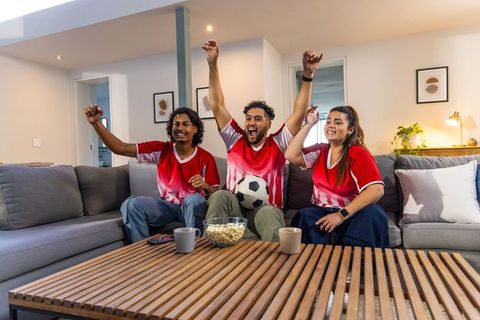 Multiethnic friends cheering at home while watching soccer match on sofa