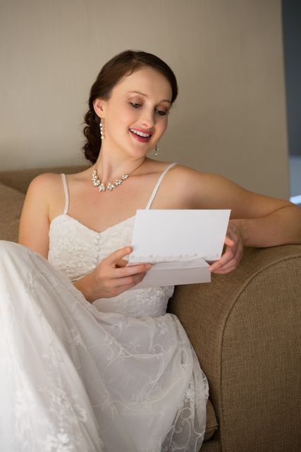 Bride in wedding dress reading a card while sitting on a sofa at home, smiling with joy. Perfect for wedding invitations, bridal magazines, wedding planning websites, and romantic greeting cards.