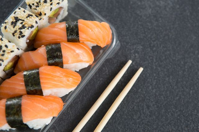 Close-up view of fresh salmon sushi rolls in a plastic container with chopsticks on a black background. Ideal for use in food blogs, restaurant menus, culinary websites, and advertisements promoting Japanese cuisine or healthy eating.