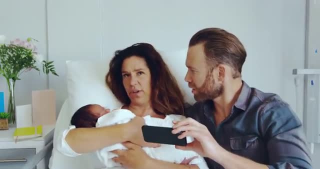 Couple holding newborn baby taking selfie in hospital ward, capturing a precious memory of new parenthood. Great for illustrating happy moments, family bond, parenthood, and maternity stories.