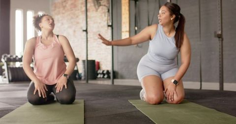 Two women kneeling on yoga mats in gym signaling during friendly workout session wearing smartwatche