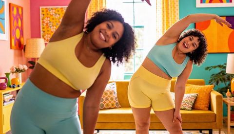 Two plus-size women stretching at home in colorful activewear on mustard sofa, midcentury living roo