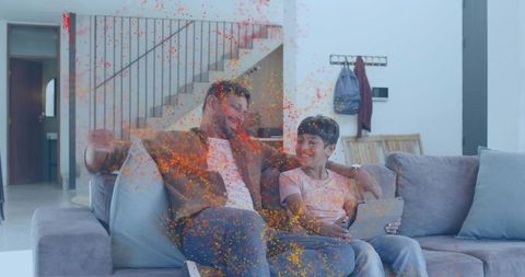 Smiling father and son sharing tablet on modern sofa celebrating with colorful confetti