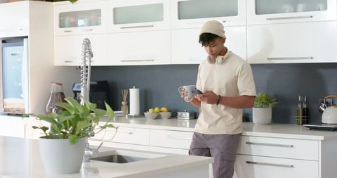 Young african man leaning on modern white kitchen counter holding mug and smartphone