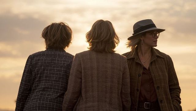 Three women standing at beach sunset wearing coats and fedora, friendship moment