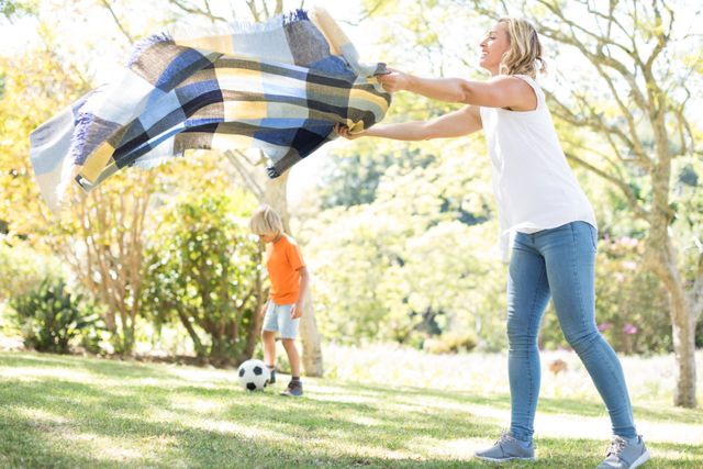 Mother spreading the picnic blanket while son playing football in background - Download Free Stock Photos Pikwizard.com