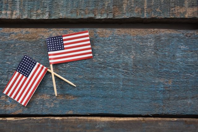 Two small American flags crossed on a rustic wooden table. Ideal for themes related to patriotism, American holidays like Independence Day, Memorial Day, and Veterans Day. Suitable for use in advertisements, social media posts, and educational materials.