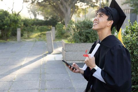 Asian graduate celebrating outdoors holding diploma and smartphone at campus golden hour