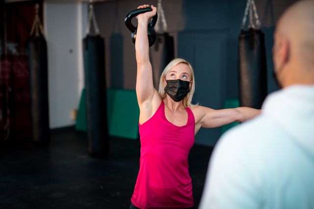 Caucasian woman wearing face mask lifting kettle ball on gym. strength and fitness cross training for boxing.