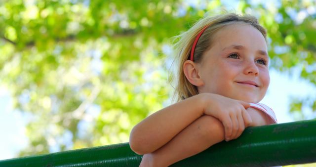 A young girl with a headband is leaning on a green bar, smiling and enjoying a sunny day outdoors with trees in the background. This can be used in campaigns focused on summer activities, children's happiness, outdoor fun, and promoting a positive, carefree lifestyle.