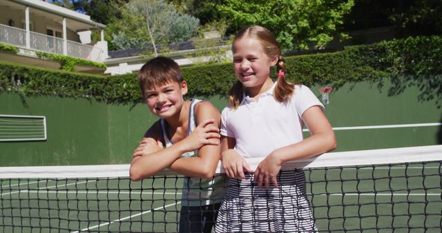 Two children leaning on tennis net, basking under sunny skies. Boy wearing tank top with arm resting on net, girl with pigtails in skirt and polo shirt. Scene ideal for illustrating youthful joy, outdoor activities, sports, sibling bond, or summer leisure.