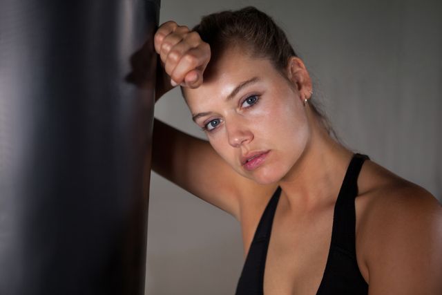 Exhausted female boxer leaning against a punching bag in a fitness studio, capturing the intensity and effort of her workout. Suitable for use in articles or promotions related to women's fitness, boxing, strength training, motivation, and athletic dedication.