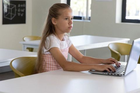 School-aged girl working on laptop in bright classroom learning digital skills and coding
