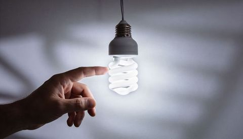 Hand touching lit spiral cfl bulb emitting soft halo glow against minimal gray wall