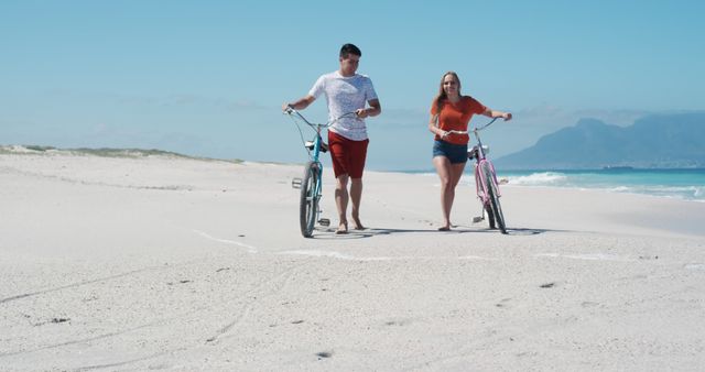 Couple walking along sandy beach pushing bicycles, clear blue sky, mountains in background. Ideal for advertisements, travel promotions, lifestyle blogs, environmental campaigns, romantic getaway themes, relaxation and leisure content.