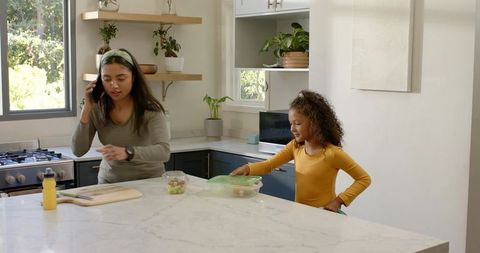 Busy mom talking on phone while daughter opening lunch container on marble kitchen island
