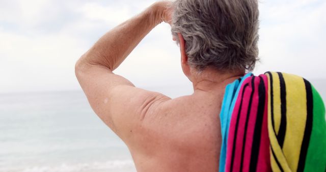 Senior man standing at the beach gazing out at the tranquil sea with a striped beach towel draped over his shoulder. Ideal for travel brochures, advertisements for retirement communities, or content featuring relaxation, serenity, and beach vacations.