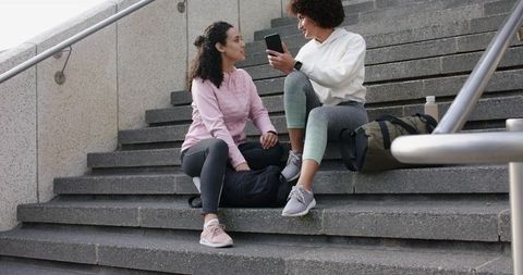 Two women sharing smartphone on campus stairs in athletic wear with backpacks and water bottle