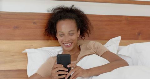 Smiling Woman Relaxing in Bed While Engaging with Smartphone