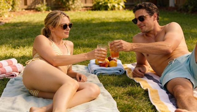 Couple toasting drinks on towels relaxing in sunny backyard wearing striped swimwear