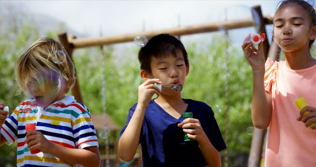 Three children are blowing bubbles outdoors on a bright sunny day, creating a lively and joyful ambiance. This image can be used for promoting children's outdoor activities, summer camps, family gatherings, and educational materials about fun and active kid's group activities.