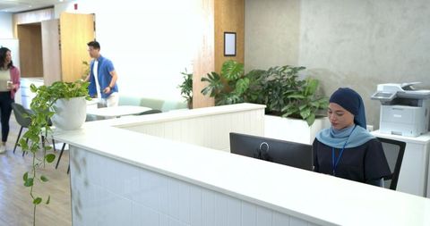 Modern office receptionist wearing headscarf at bright front desk with plants and colleagues walking
