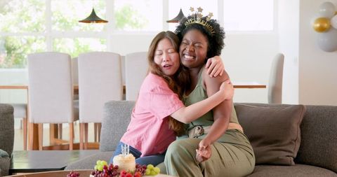 Two women celebrate a birthday while sitting on a sofa, exchanging a warm hug. Surrounded by fruit and cake, they embody joy and friendship. Perfect for illustrating diverse relationships, celebrations, and joy at home. Ideal for use in articles or campaigns about friendship, diversity, or personal celebrations.