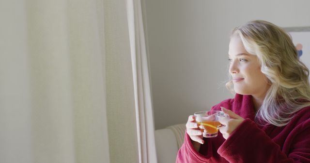 Woman in red bathrobe enjoying tea by a window, capturing a moment of calm and relaxation. Ideal for content on self-care, lifestyle blogs, morning routines, health and wellness, and advertisements promoting relaxation products or home living.