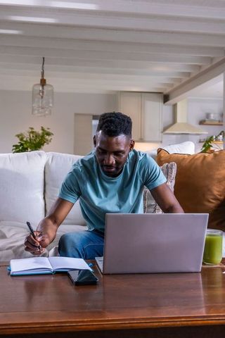 Man working from home on sofa writing notes while using laptop for remote productivity