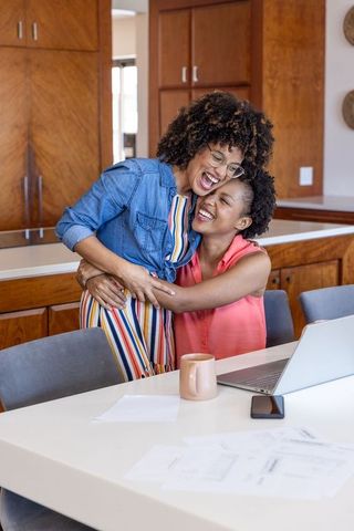 Laughing diverse female couple hugging at kitchen table while working remotely on laptop